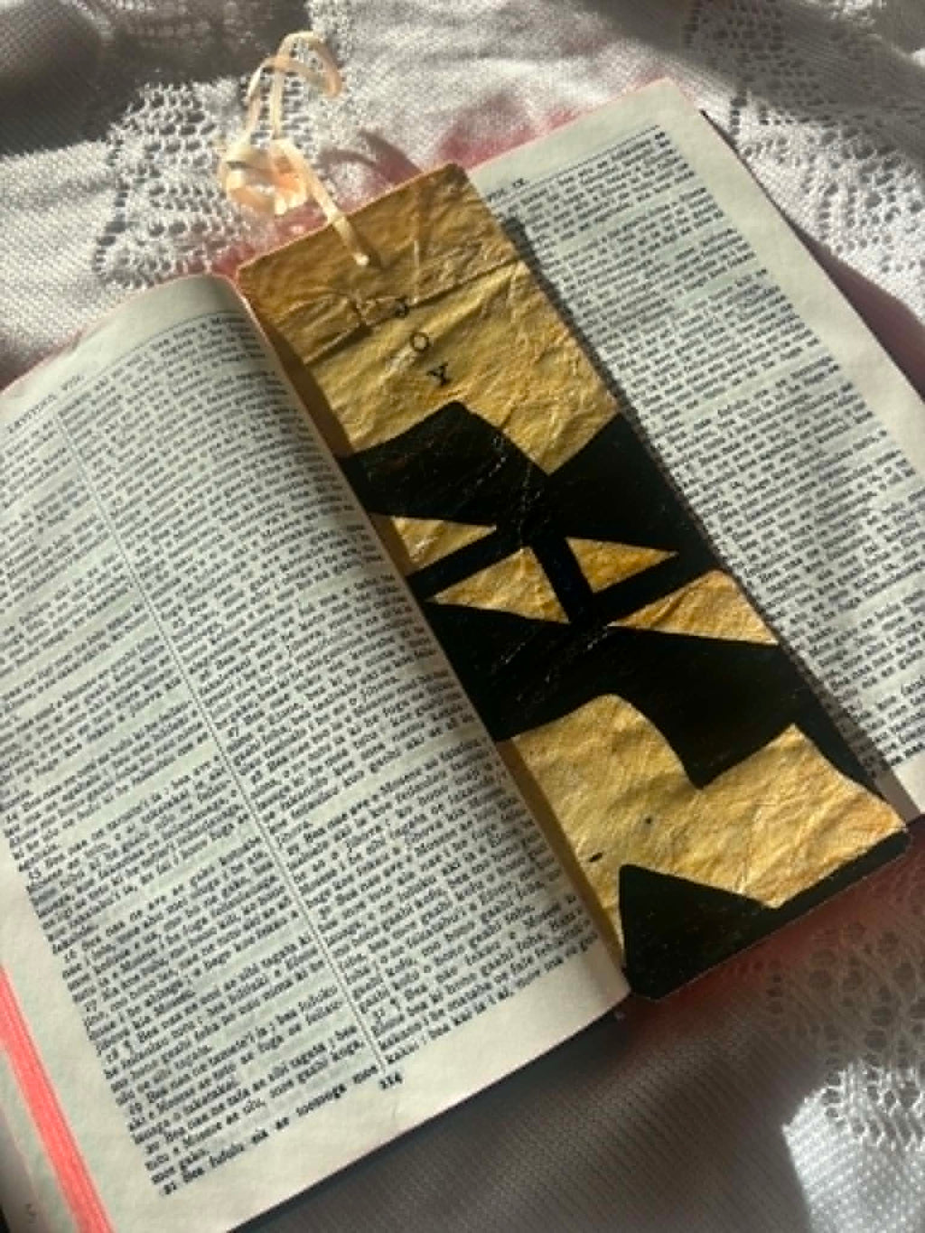 Gold bookmark with black letter wordmark and tassel, lying on open scripture text